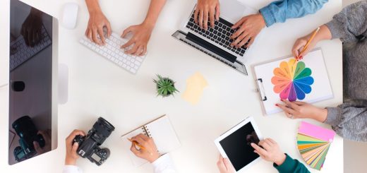 A top view of four people sitting around a table with laptops, papers, drinks, and notebooks in a bright office.