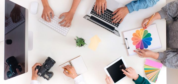 A top view of four people sitting around a table with laptops, papers, drinks, and notebooks in a bright office.