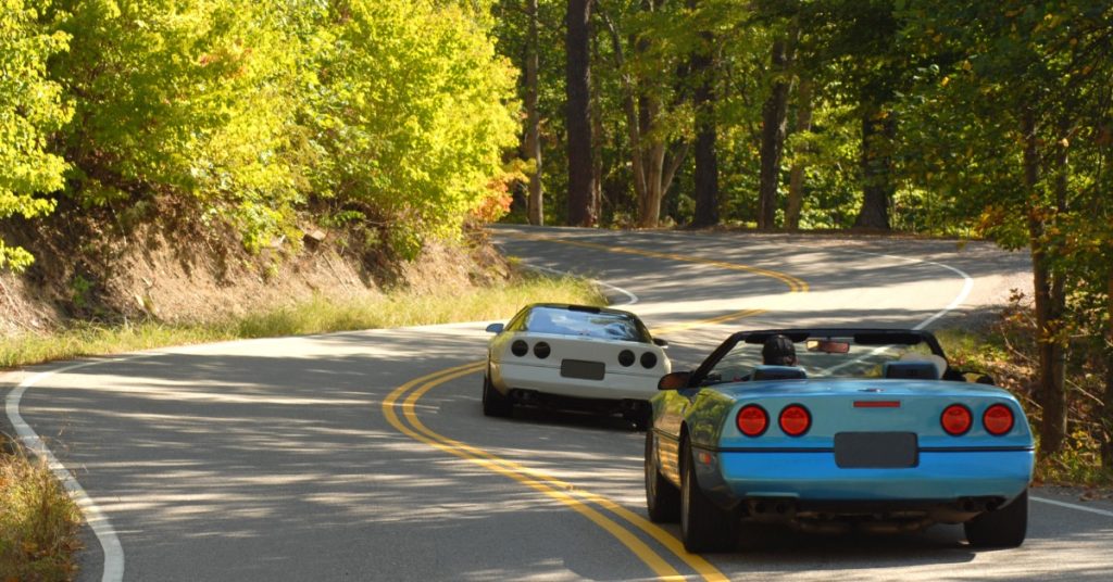 A rear view of two sports cars, one blue with the top down and one white with the top on, driving on a two-lane forest road.