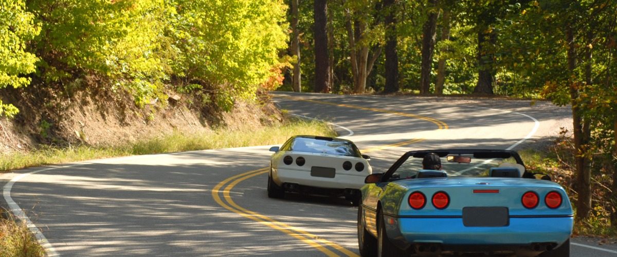 A rear view of two sports cars, one blue with the top down and one white with the top on, driving on a two-lane forest road.