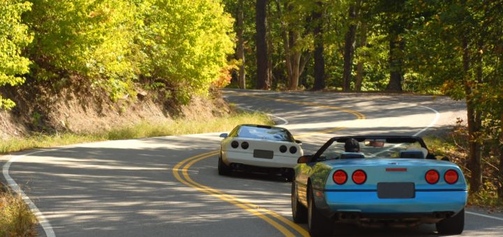 A rear view of two sports cars, one blue with the top down and one white with the top on, driving on a two-lane forest road.