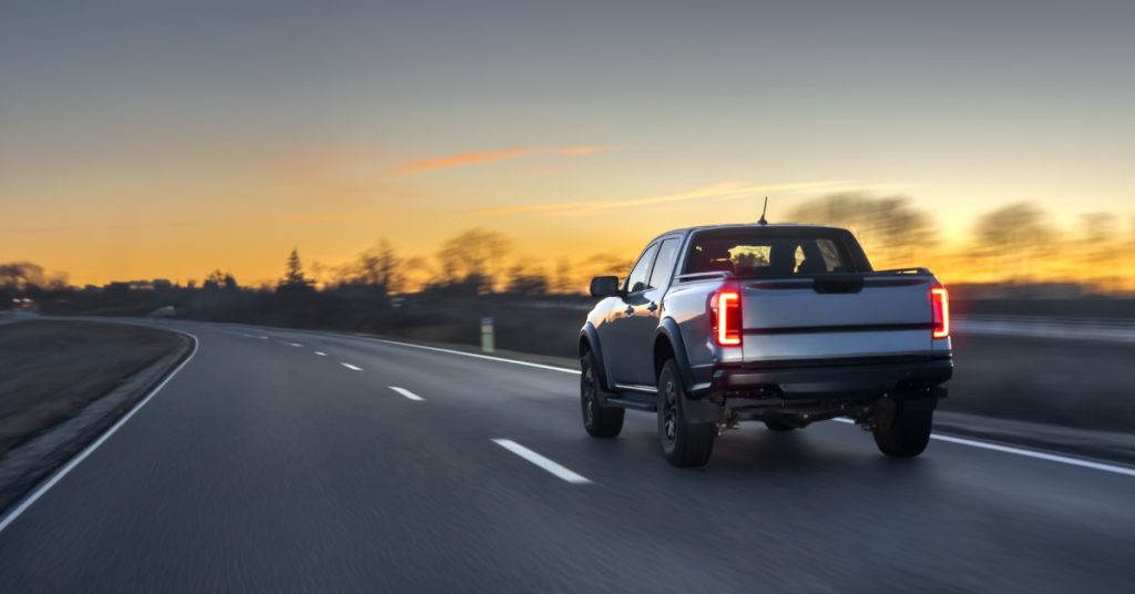 A grey pickup truck with the brake lights on, driving on a concrete road with a blurred background during a sunset.