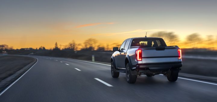A grey pickup truck with the brake lights on, driving on a concrete road with a blurred background during a sunset.