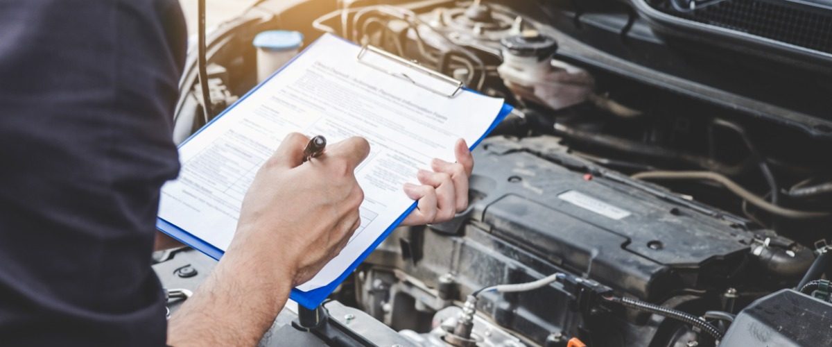 A person is holding a clipboard that has a sheet of paper on it. They are doing this while looking at a car engine.