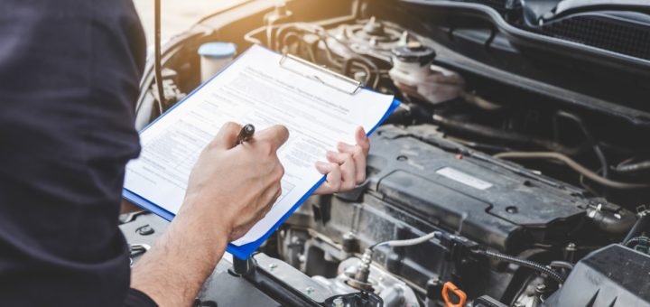 A person is holding a clipboard that has a sheet of paper on it. They are doing this while looking at a car engine.