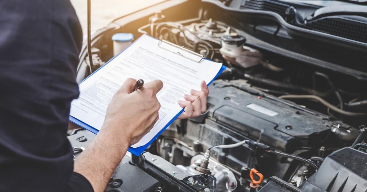A person is holding a clipboard that has a sheet of paper on it. They are doing this while looking at a car engine.