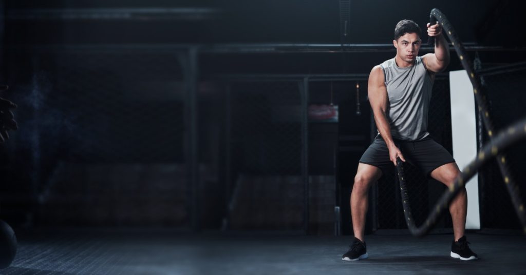 A young man is swinging battle ropes in a dimly lit gym while a person prepares to grab a kettlebell in the shadows.