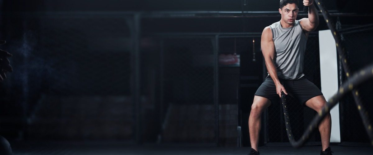 A young man is swinging battle ropes in a dimly lit gym while a person prepares to grab a kettlebell in the shadows.