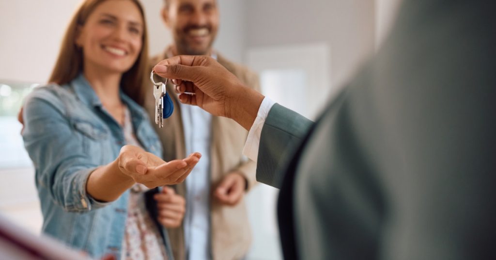 A close-up of the hand of a person holding a set of keys over the hand of a woman standing next to a man in a room.