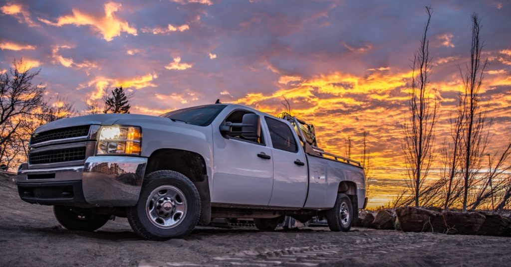 A white heavy-duty diesel pickup truck parked on rough terrain beneath a dramatic orange and purple sunset sky.
