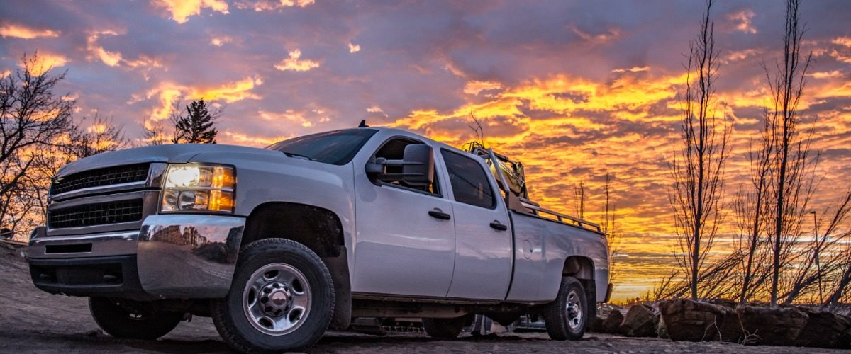 A white heavy-duty diesel pickup truck parked on rough terrain beneath a dramatic orange and purple sunset sky.