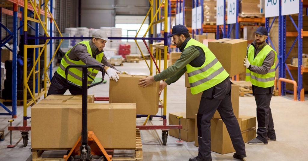 Three men are in a warehouse. Two of the men are stacking boxes on a pallet while the third is handing the two men more boxes.