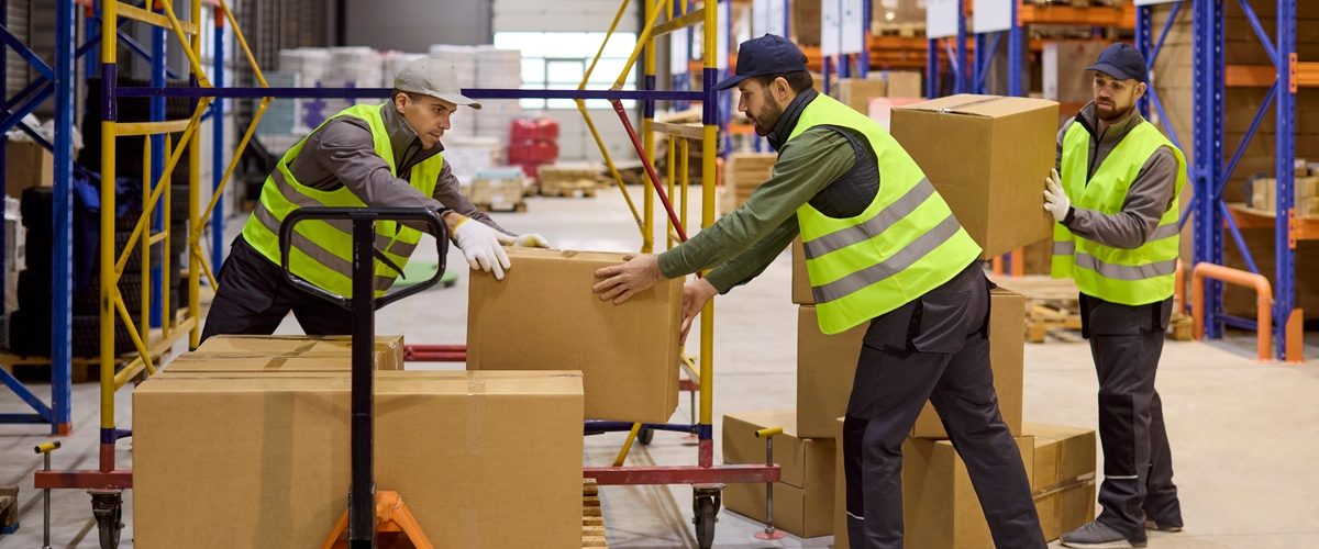 Three men are in a warehouse. Two of the men are stacking boxes on a pallet while the third is handing the two men more boxes.