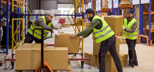 Three men are in a warehouse. Two of the men are stacking boxes on a pallet while the third is handing the two men more boxes.