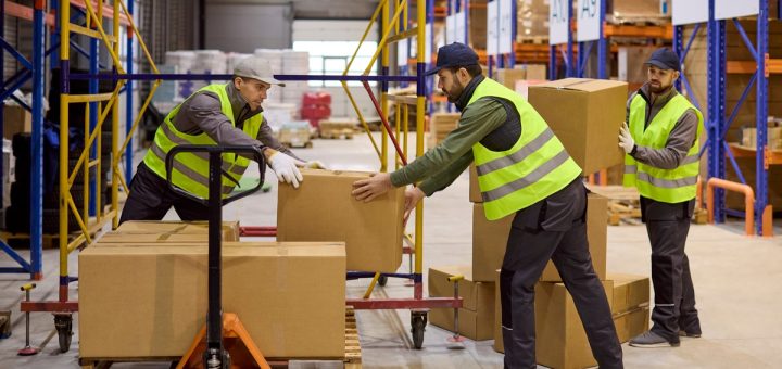 Three men are in a warehouse. Two of the men are stacking boxes on a pallet while the third is handing the two men more boxes.