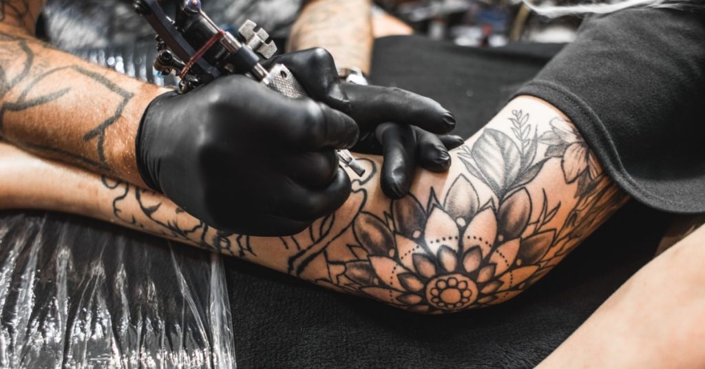 A close-up of an arm with multiple black ink tattoos on a table, with a hand holding an ink gun close to the arm.