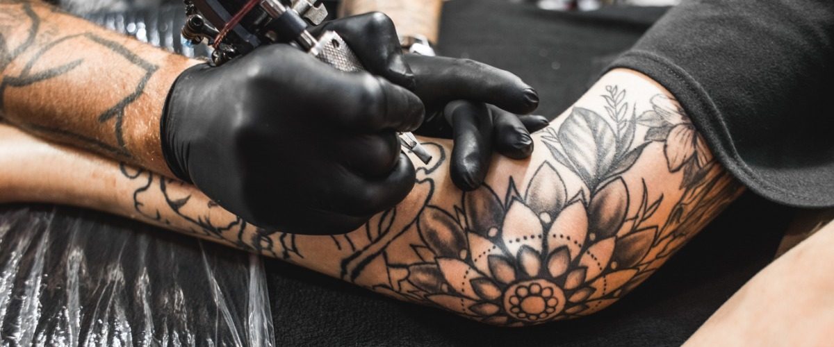 A close-up of an arm with multiple black ink tattoos on a table, with a hand holding an ink gun close to the arm.
