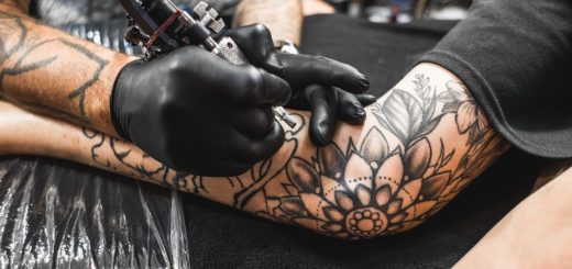 A close-up of an arm with multiple black ink tattoos on a table, with a hand holding an ink gun close to the arm.