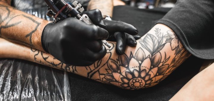 A close-up of an arm with multiple black ink tattoos on a table, with a hand holding an ink gun close to the arm.