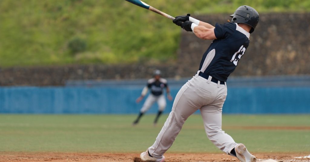 A rear view of a baseball batter with a black helmet, black jersey, and grey pants swinging at the plate.