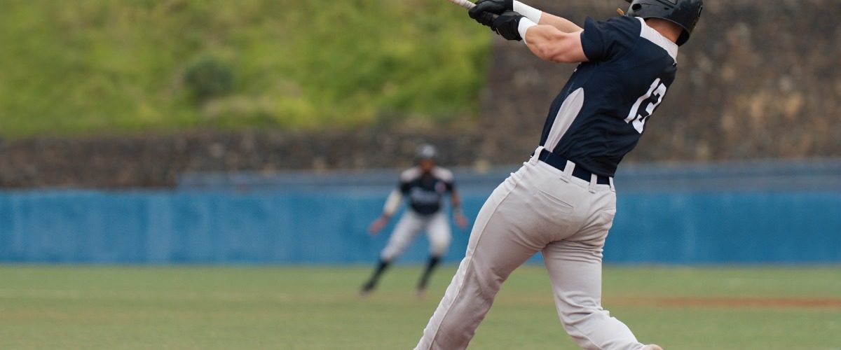 A rear view of a baseball batter with a black helmet, black jersey, and grey pants swinging at the plate.