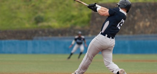 A rear view of a baseball batter with a black helmet, black jersey, and grey pants swinging at the plate.
