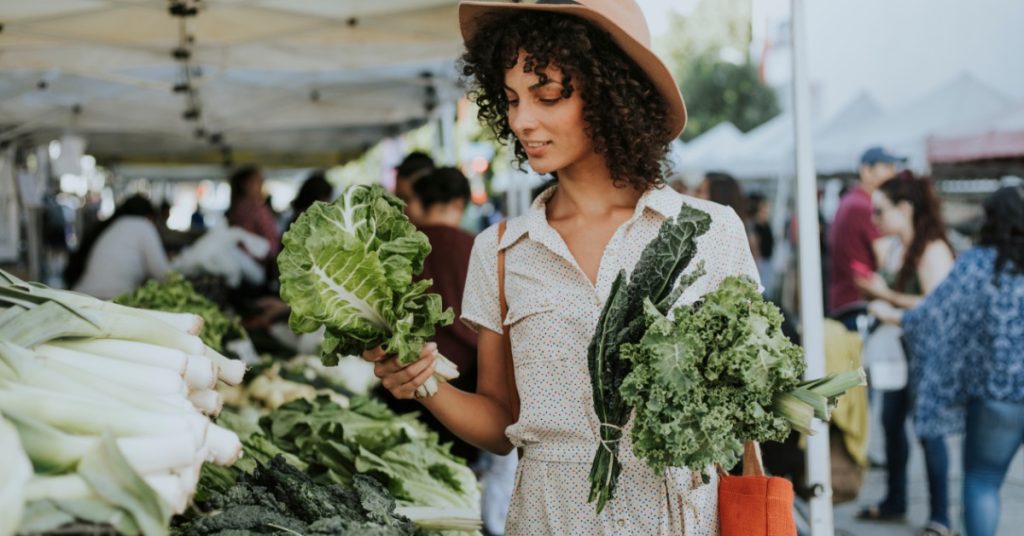 Shopper wearing a brown hat holds handfuls of kale outside at a crowded farmers market on a sunny day.