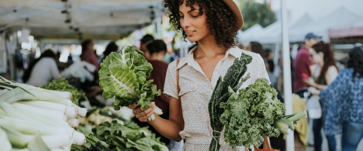 Shopper wearing a brown hat holds handfuls of kale outside at a crowded farmers market on a sunny day.