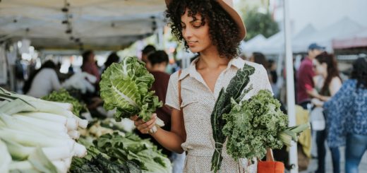 Shopper wearing a brown hat holds handfuls of kale outside at a crowded farmers market on a sunny day.