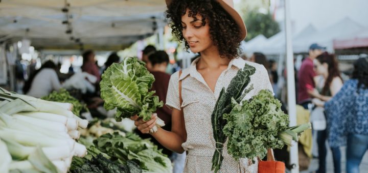 Shopper wearing a brown hat holds handfuls of kale outside at a crowded farmers market on a sunny day.