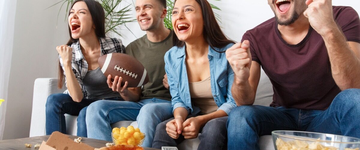 A group of four people sit on the couch together eating pizza as they tune into a football game on the TV.