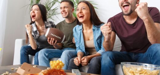 A group of four people sit on the couch together eating pizza as they tune into a football game on the TV.