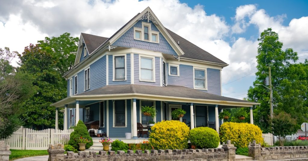 A large two-story house with white siding, green shutters, a front porch, shrubs, and a lawn under a blue sky.