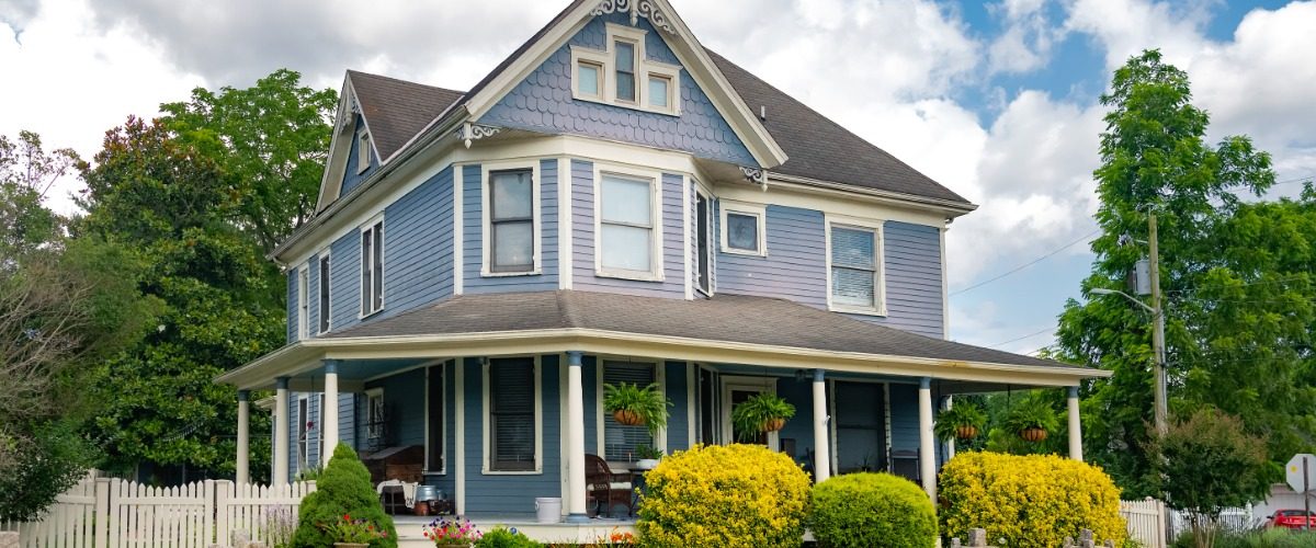 A large two-story house with white siding, green shutters, a front porch, shrubs, and a lawn under a blue sky.
