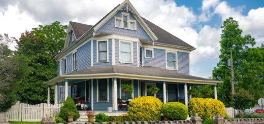 A large two-story house with white siding, green shutters, a front porch, shrubs, and a lawn under a blue sky.