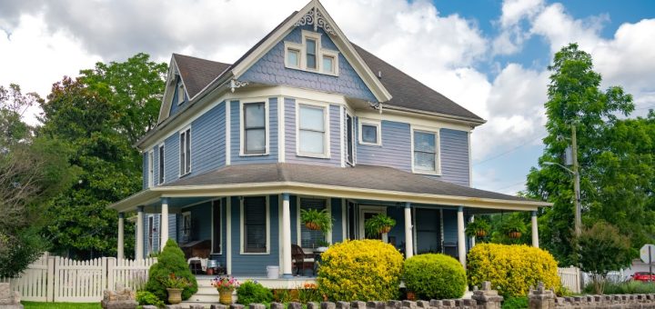 A large two-story house with white siding, green shutters, a front porch, shrubs, and a lawn under a blue sky.