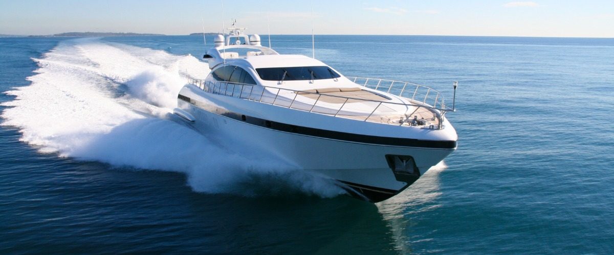 Large white luxury yacht speeds across calm blue ocean water, creating a wide trail of white spray behind it.