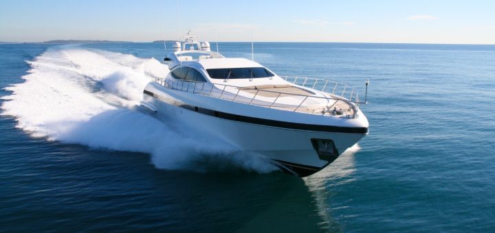 Large white luxury yacht speeds across calm blue ocean water, creating a wide trail of white spray behind it.