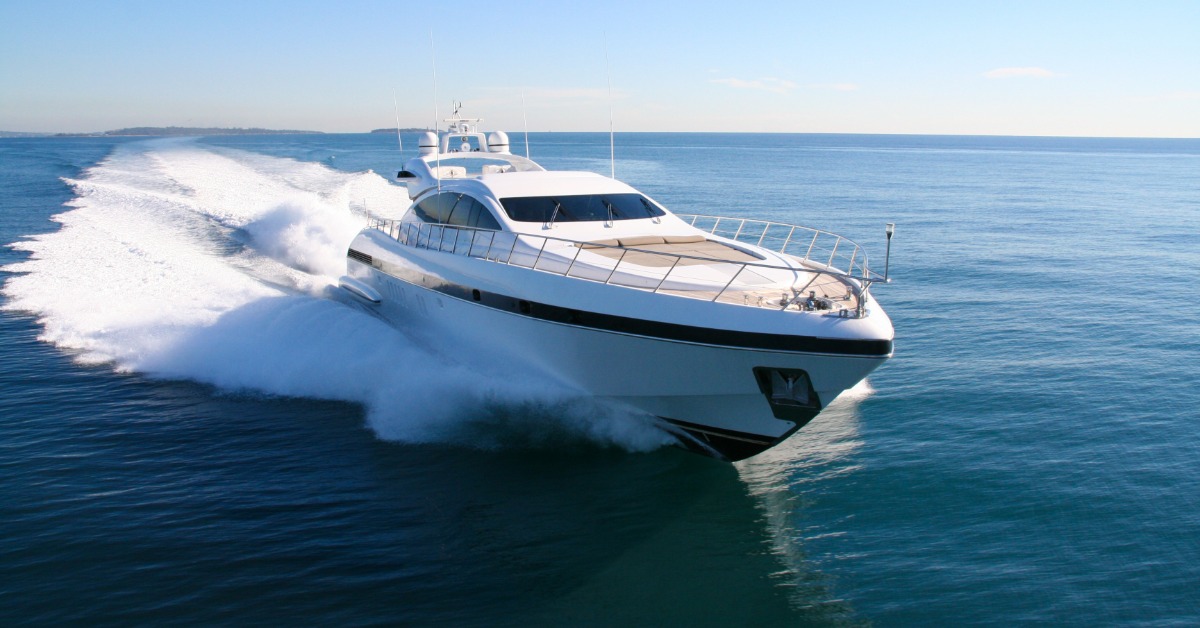 Large white luxury yacht speeds across calm blue ocean water, creating a wide trail of white spray behind it.