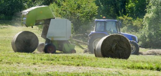 A green hay baler attached to a blue tractor outside on a sunny day. Two round hay bales lay on the ground nearby.
