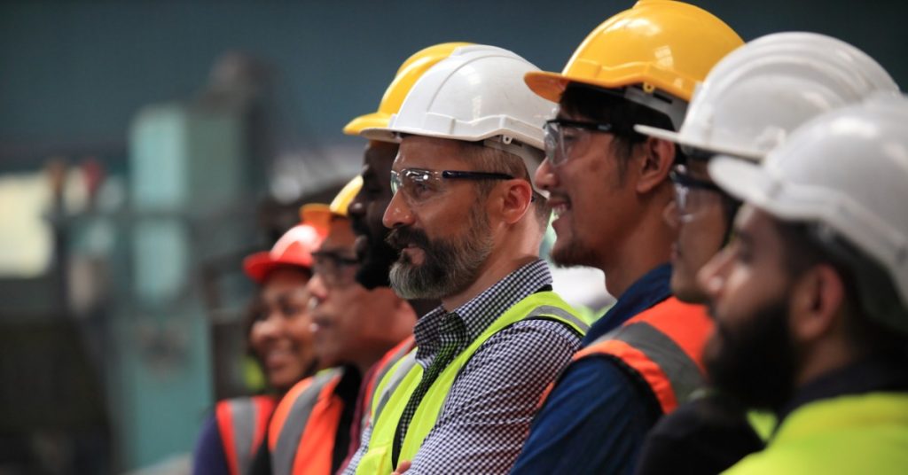 Several people are lined up. They are all wearing hardhats, protective eyewear, and high-visibility vests.