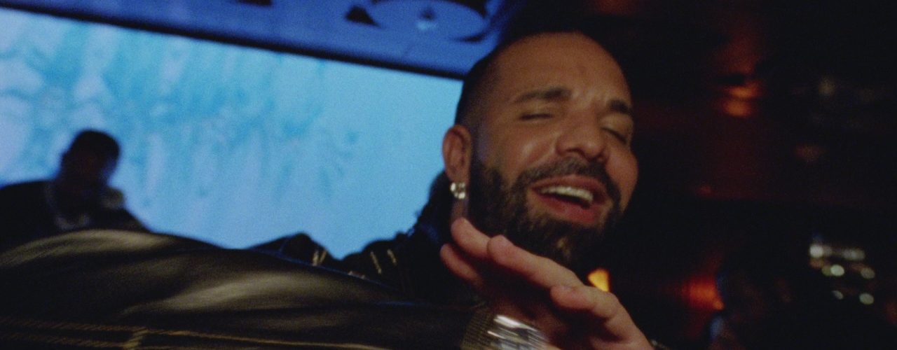 Drake smiles with a hand raised in a dim lounge in a still from his new music video
