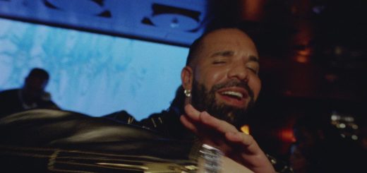Drake smiles with a hand raised in a dim lounge in a still from his new music video