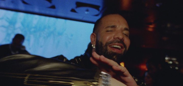 Drake smiles with a hand raised in a dim lounge in a still from his new music video
