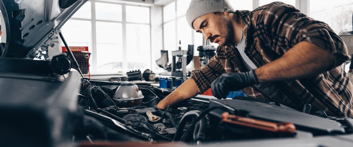 A man with a knit cap on his head is leaning over a car’s engine block. There are windows in the background.