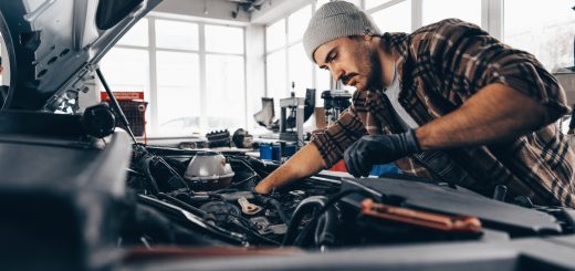A man with a knit cap on his head is leaning over a car’s engine block. There are windows in the background.