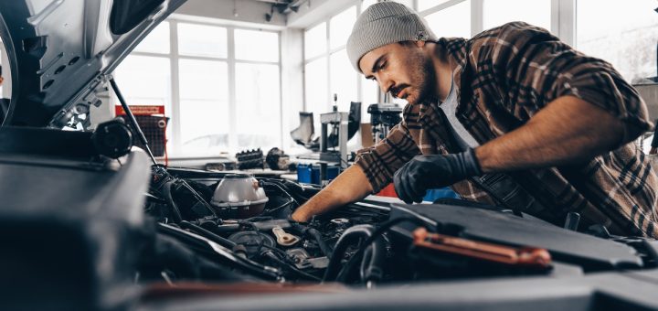 A man with a knit cap on his head is leaning over a car’s engine block. There are windows in the background.