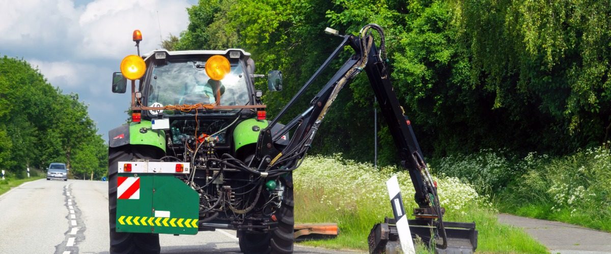 A large, green machine is using a boom mower to cut the grass of the side of the road. There is a car on the road.
