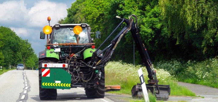 A large, green machine is using a boom mower to cut the grass of the side of the road. There is a car on the road.