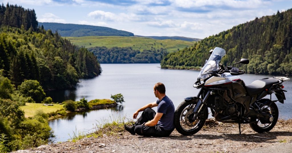 A man wearing black leather pants sitting on the ground next to a black adventure motorcycle on the edge of a cliff.